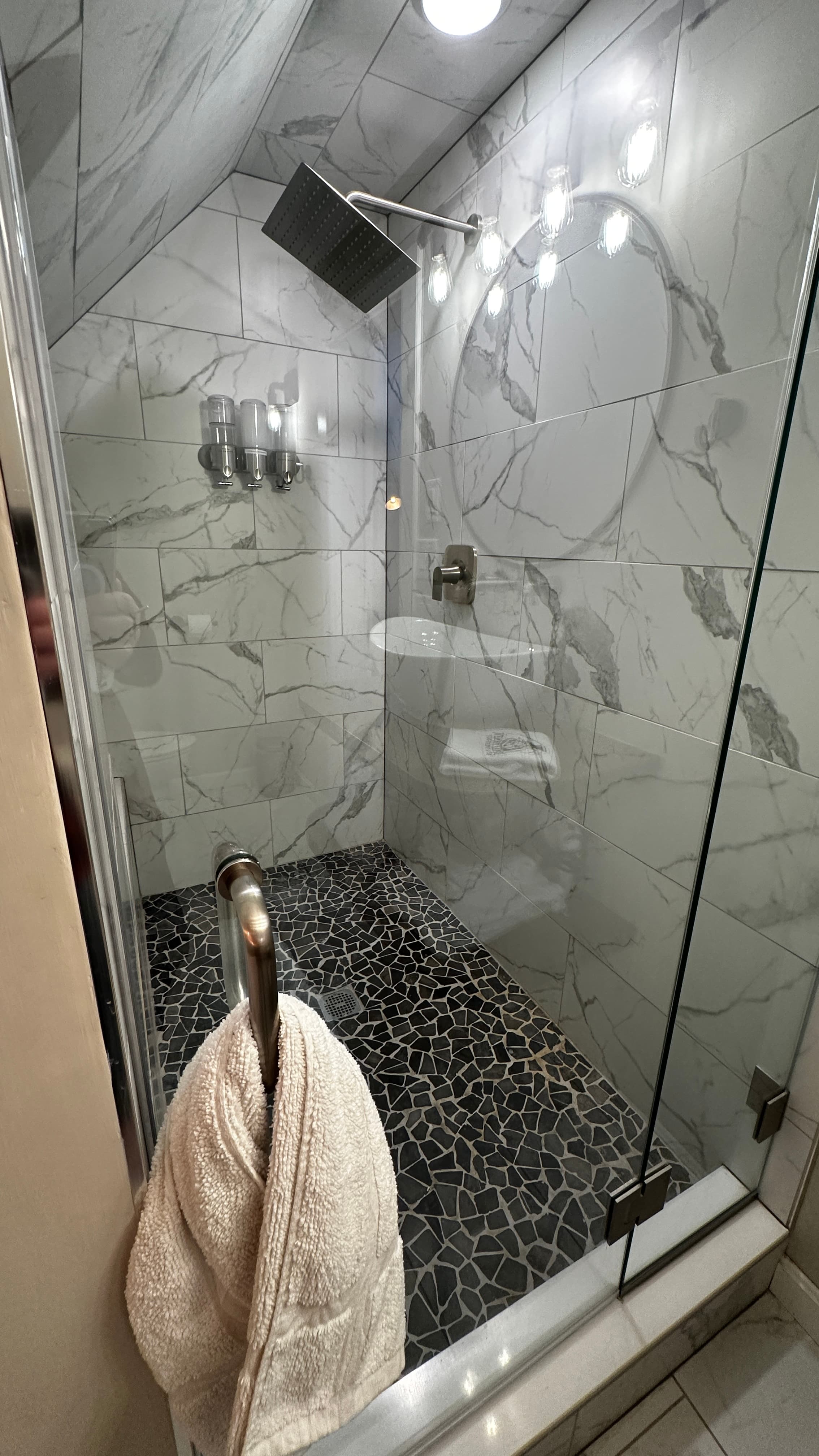 A close-up of a modern walk-in shower featuring a glass door and white marble-patterned wall tiles. The shower includes a dark square rainfall showerhead, a pebble-style floor, and a white towel draped over the handle in the foreground.