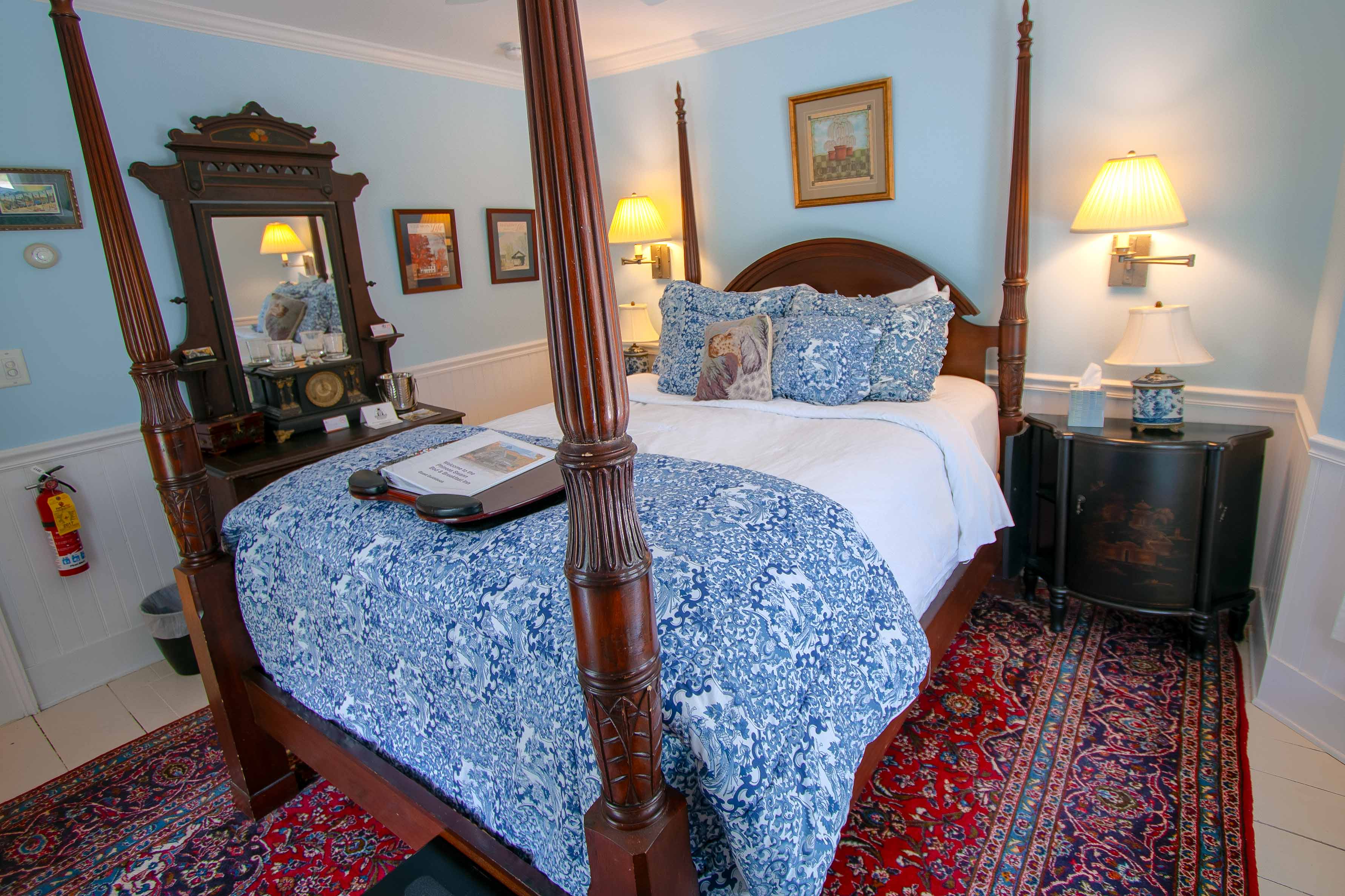 A four-poster bed with blue floral linens stands in a bright guest room with light blue walls and white wainscoting. An antique dark wood dresser with a large ornate mirror and a red patterned rug complete the traditional decor.