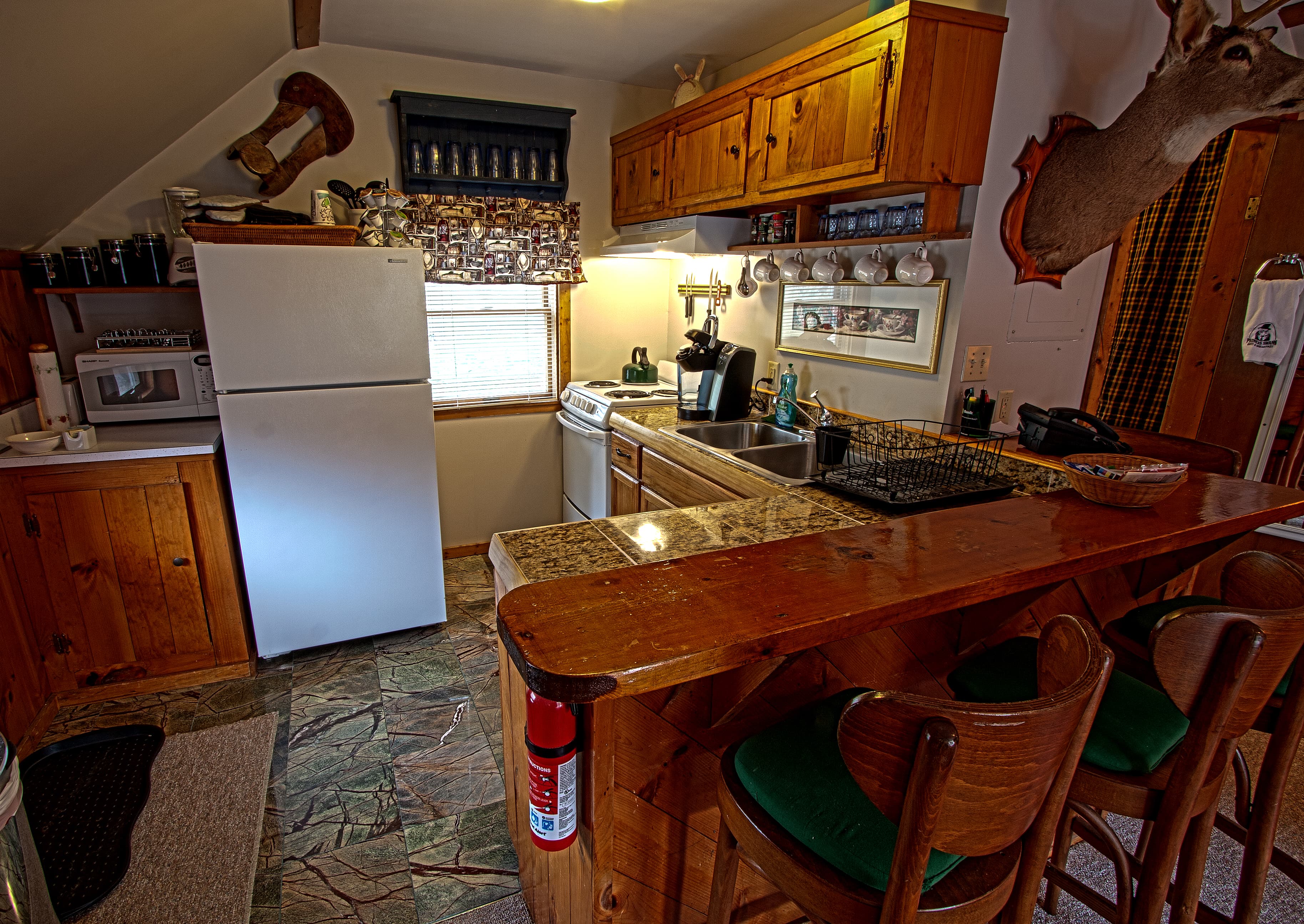 A small, rustic kitchen with wooden cabinets and a white refrigerator. A wooden breakfast bar with green-cushioned stools sits in the foreground, and a mounted deer head is visible on the wall to the right.