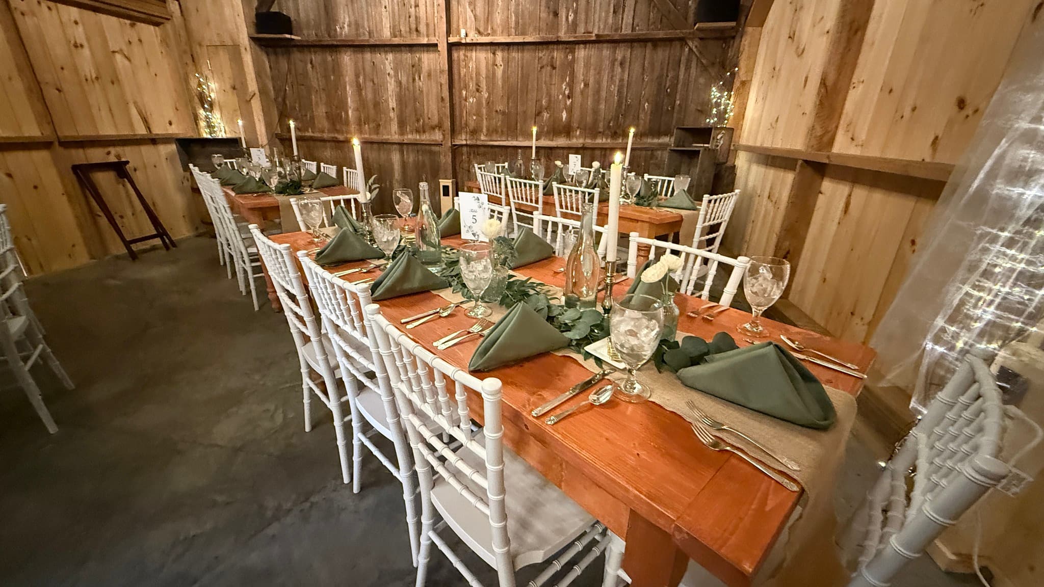 A rustic dining setup with wooden tables, white chairs, and green napkins, arranged elegantly in a barn interior.