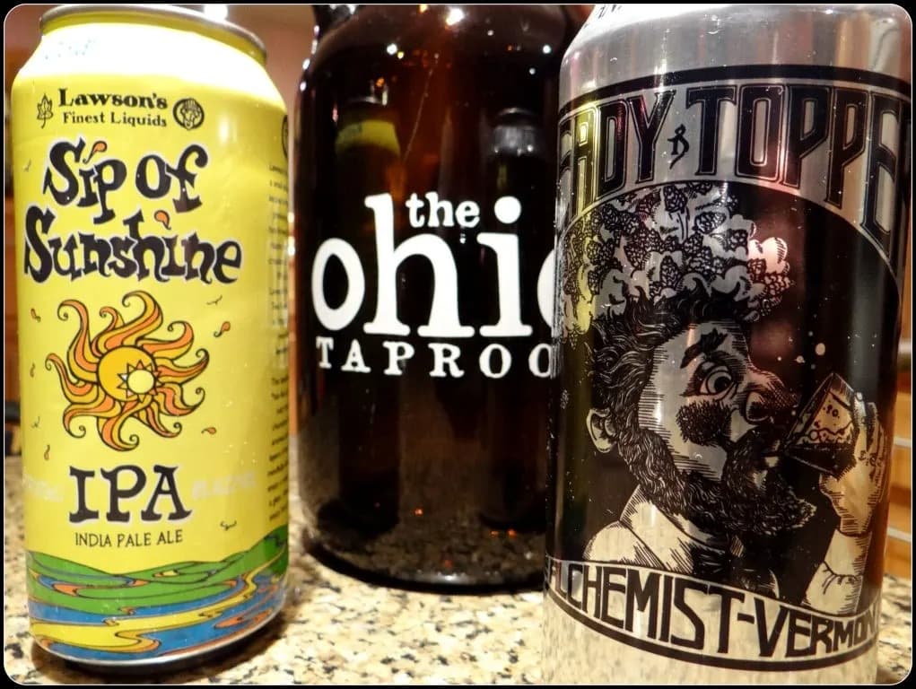 A can of "Sip of Sunshine" IPA, a growler from "The Ohio Taproom," and a can of "Heady Topper" beer are displayed together on a countertop.