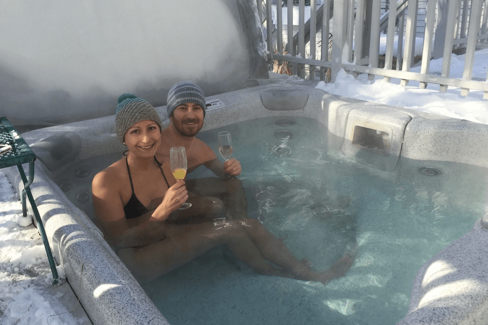 A couple relaxes in a hot tub surrounded by snow, each holding a drink.