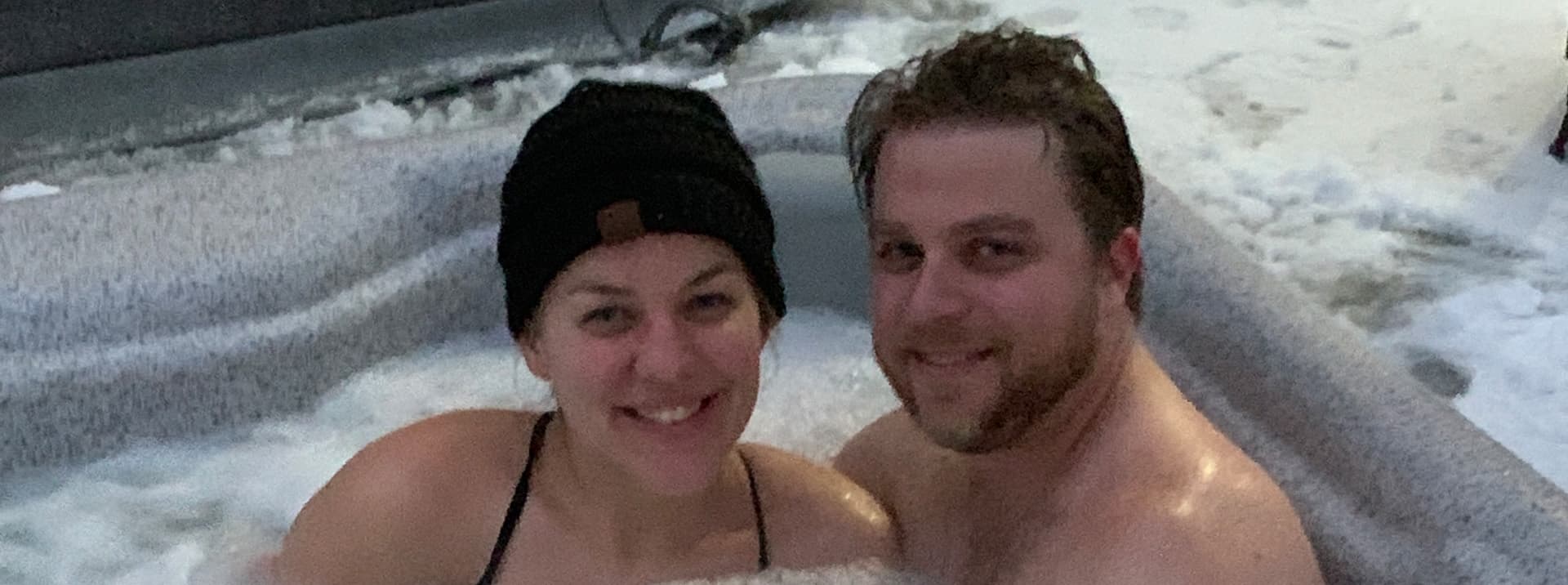 A smiling couple relaxes in a hot tub surrounded by snow.