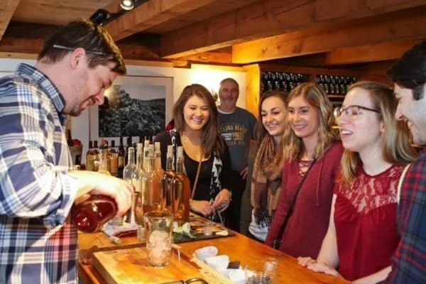 A bartender pours a drink while a group of smiling friends watches with curiosity in a cozy bar setting.