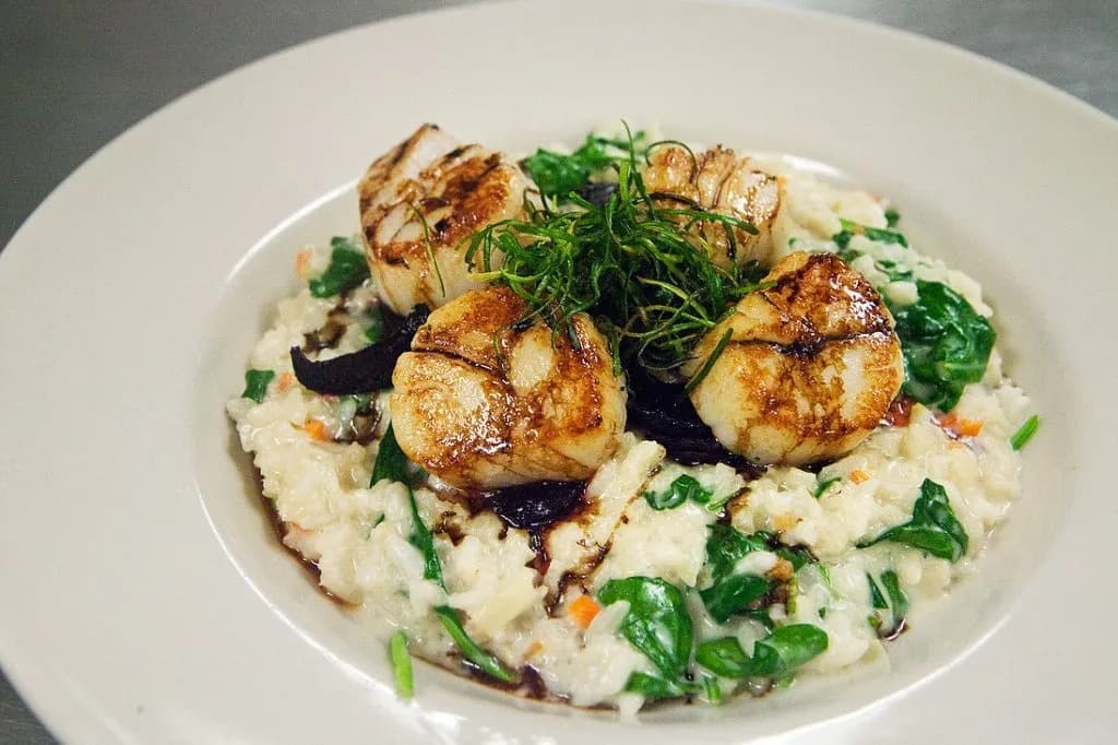 A plate of creamy risotto topped with three seared scallops and garnished with leafy greens.