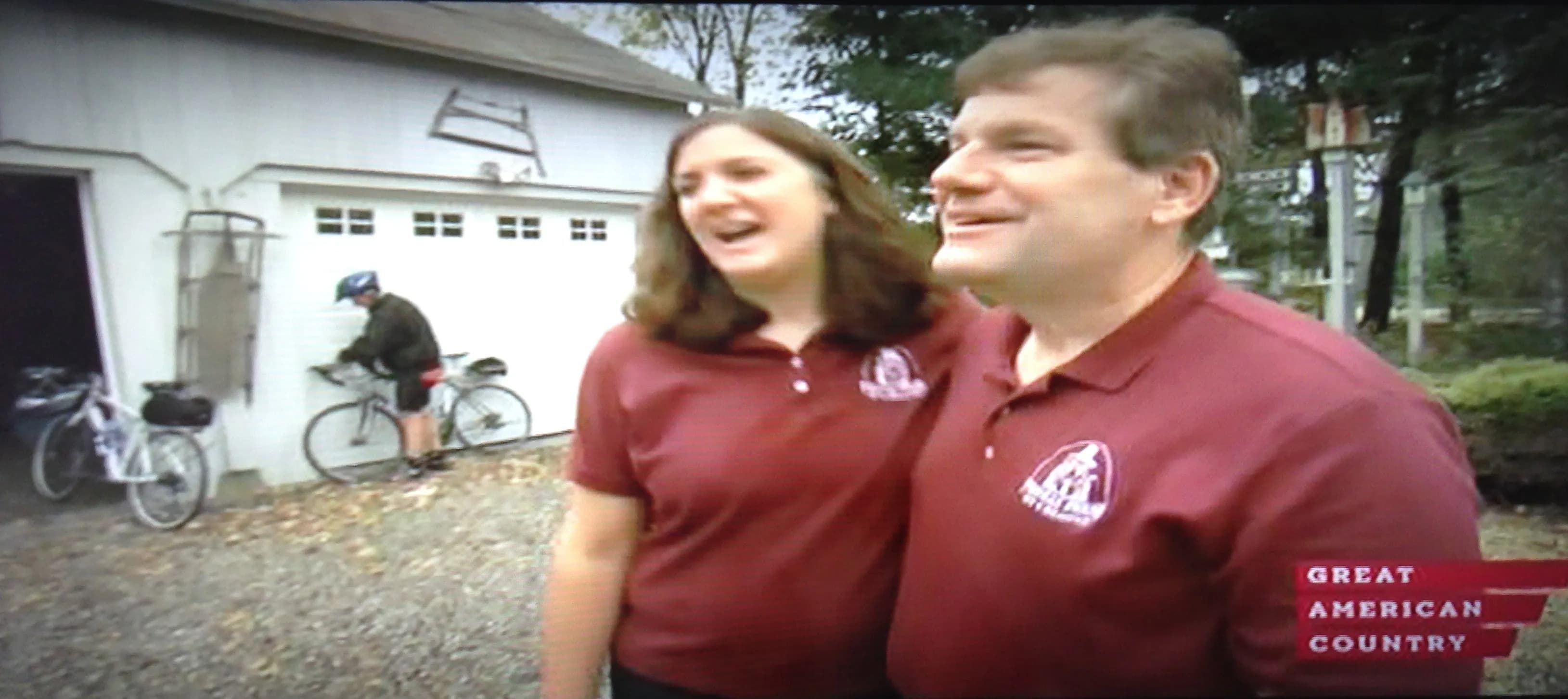 A smiling couple stands together outside a garage, while a person prepares to ride a bicycle in the background.