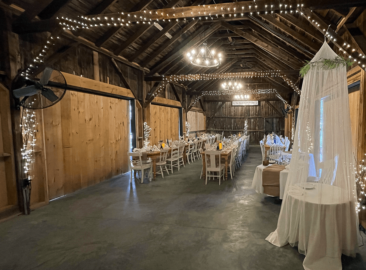 A rustic barn interior decorated with lights, featuring long dining tables set for an event.