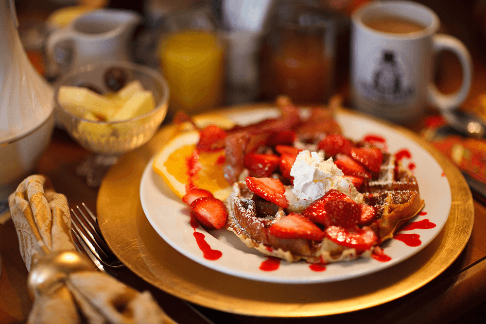 A golden-brown waffle topped with strawberries, whipped cream, and a drizzle of syrup, surrounded by fruit and beverages.