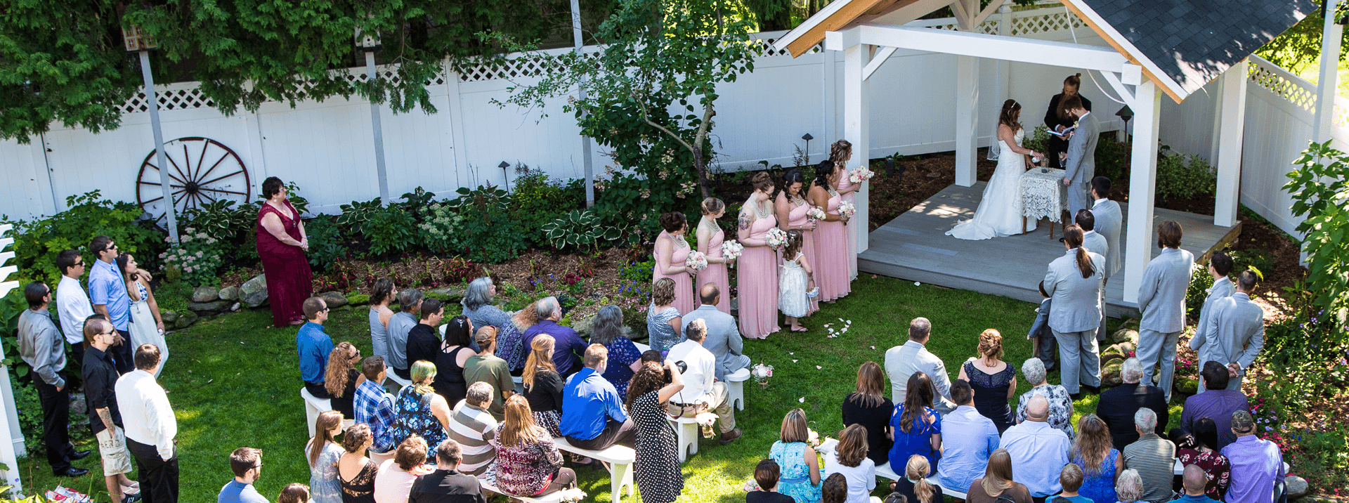 A wedding ceremony takes place outdoors under a white gazebo, surrounded by guests in a garden setting.