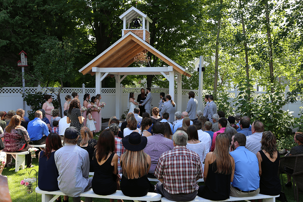 A wedding ceremony takes place outdoors under a gazebo, surrounded by guests seated on benches.